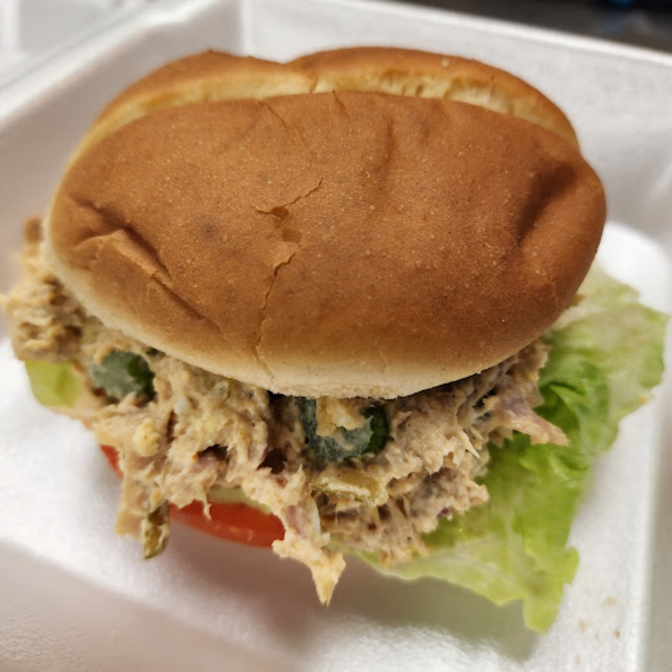 A tuna salad sandwich on a bun with lettuce and tomato in a white takeout container.